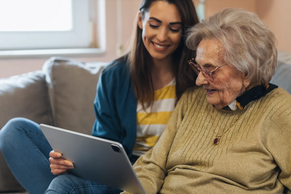 An adult daughter sits beside her elderly mother on a couch, smiling as they look at a tablet together.
