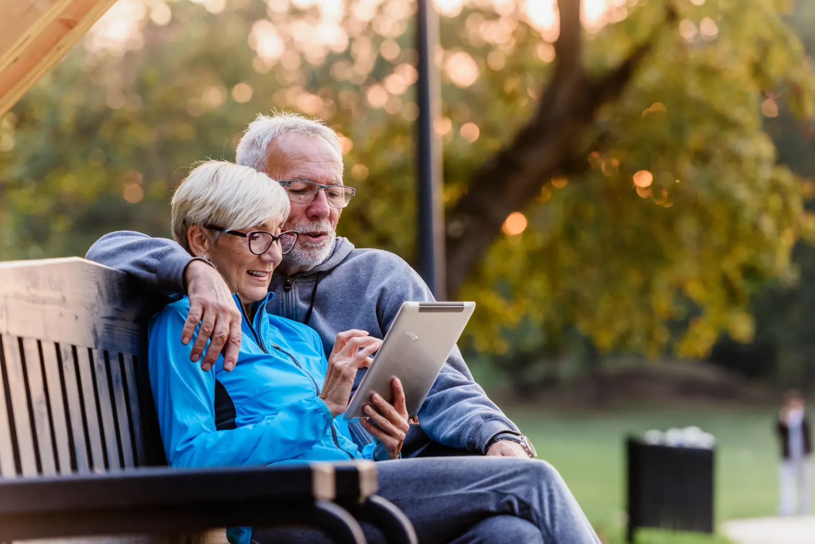 A retired couple sit together on a park bench, reviewing something on a tablet in warm evening light.